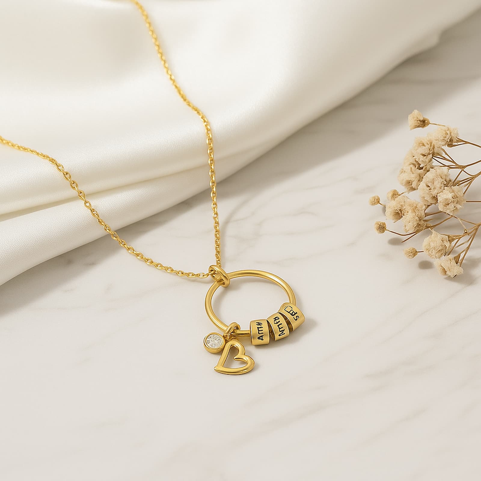 Close-up on the gold personalized circle necklace against a silky background, clearly showing the engraved name beads and heart charm.