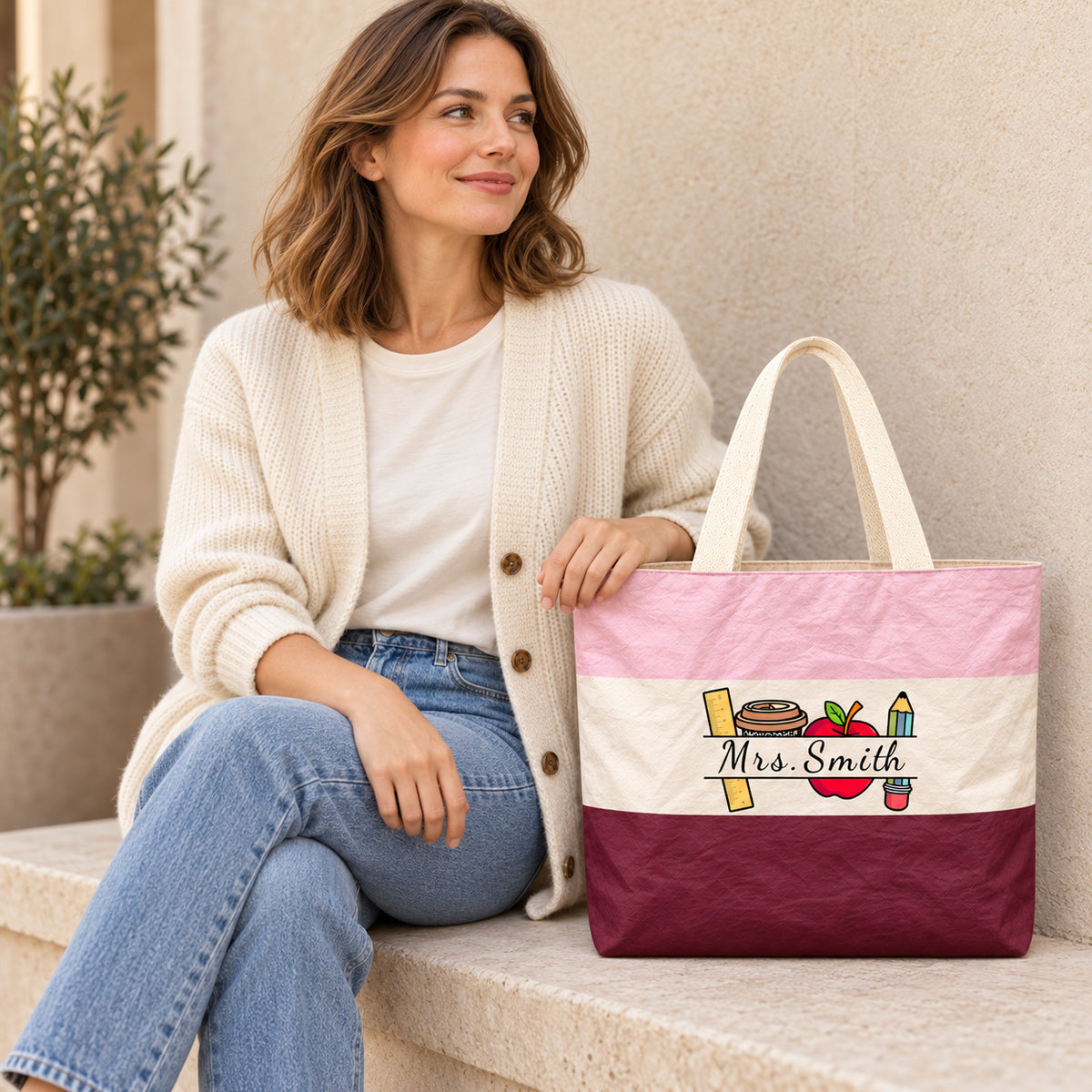 Woman seated beside a colorblock teacher tote showing the custom name Mrs. Smith and school supplies artwork.