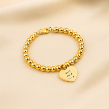 Gold bead ball strand bracelet with a custom heart charm, displayed on a soft, light-colored fabric backdrop.