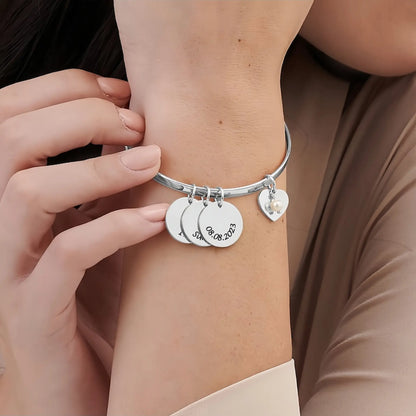 A woman wearing a silver bangle bracelet with multiple engraved circular charms, a heart-shaped charm, and a small pearl accent on her wrist.