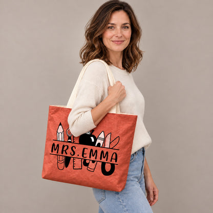 Personalized teacher tote with school supplies artwork and custom name MRS.EMMA, shoulder side view view.